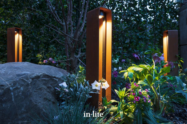 Atmospheric lighting photography by in-lite. ACE HIGH Corten 12v outdoor bollard luminaires positioned between border rockery and plants illuminating with targeted light beams downwards.