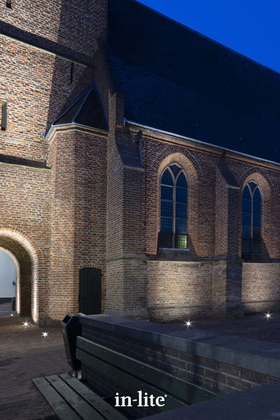 Atmospheric lighting by in-lite. FLUX 12v outdoor ground recessed light fixtures illuminating structural stone elements and brick wall facades of an architectural town building.