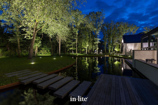 A wide evening shot of a high-value estate garden centred around a dark reflecting pool. High-output in-lite BIG NERO ground fixtures are flush-mounted into the lawn, directly uplighting a line of mature tree canopies along the far bank to highlight the site's natural capital. The illuminated trees reflect sharply in the still water.