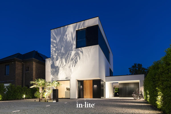 Atmospheric lighting by in-lite. BIG SCOPE 12v fixtures featured in a photograph taken at night, during the blue hour, of a modern white cubic house. Warm landscape lighting illuminates palm trees and a gravel driveway in the foreground. A distinct shadow of a palm frond is cast onto the upper wall of the house. The front entrance features a wooden door and large glass panels, with an adjacent black garage area and a bicycle. An "in-lite" logo is present at the bottom of the gravel.