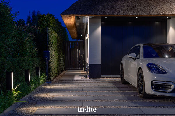 Atmospheric lighting by in-lite. EVO LOW DARK 12v outdoor architectural bollards, positioned in border, illuminating paved driveway and hybrid Porsche with diffused light in modern landscape.