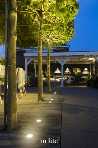 Atmospheric lighting by in-lite. FLUX 12v outdoor ground recessed light fixtures illuminating pleached trees around the perimeter space of a deck with dining table and seating.