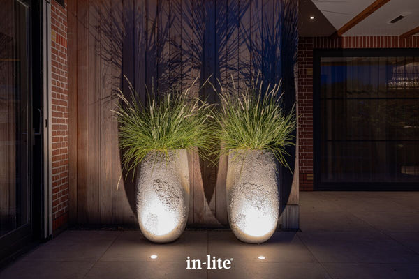 An evening shot of a modern patio featuring two tall, textured stone planters filled with ornamental grasses. Flush-mounted in-lite LUNA recessed ground lights installed in the premium paving brightly uplight the planters, highlighting their rough texture and casting dramatic, architectural shadows of the foliage onto the vertical timber-clad wall behind. The setting is framed by contemporary brickwork and large glass doors.