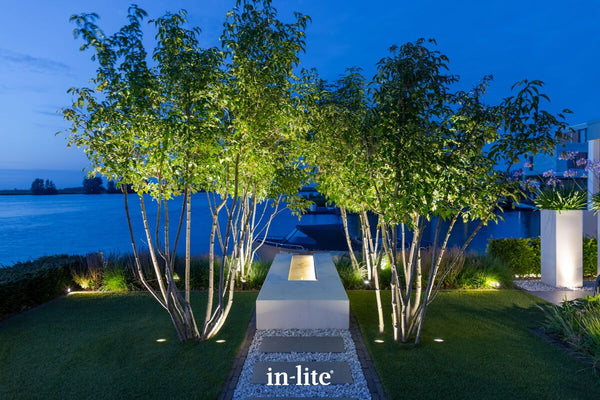 A high-value waterfront landscape at dusk. Two symmetrical multi-stemmed trees are sharply uplit by in-lite NERO recessed ground lights, framing a modern, illuminated rectangular focal point on a gravel and stone path. The flush-mounted, low-voltage fixtures deliver targeted canopy illumination while maintaining a clean lawn aesthetic against the backdrop of a lake and moored boat.