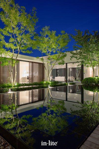 Atmospheric lighting by in-lite. SCOPE 12v outdoor spotlight fixtures illuminating architecturally favoured trees around an inifnity pool in the grounds of a modern architecture property. The landscape lighting and the reflection in the pool are incredible, creating a beautiful nighttime shot with dramatic effects in a balanced and landscaped grounds of a premium modern property.