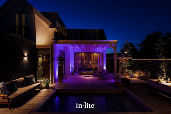 Atmospheric lighting by in-lite. SMART FLUX TONE 12v outdoor ground recessed vertical lighting fixtures washing exterior walls of property with a vibrant colour within a pergola covered patio lounge space outdoors.