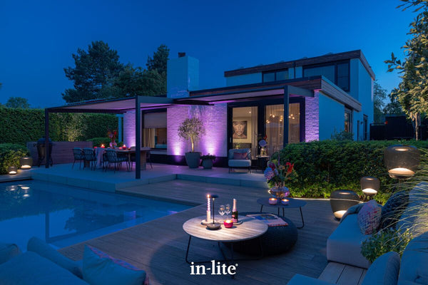 Atmospheric lighting by in-lite. SMART FLUX TONE 12v outdoor ground recessed vertical lighting fixtures illuminating a patio transformed into a lounge through deep purple hues highlighting the brickwork, which contrasts sharply with the warm, inviting amber glow seen through the home’s glass doors.