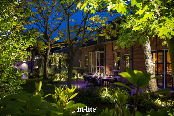 A luxurious, softly illuminated evening garden scene featuring a brick property and an elegantly set outdoor dining patio. Warm, low-voltage in-lite landscape lighting strategically uplights the canopies of mature trees and highlights low-level ferns and planting beds, showcasing high-end biophilic design and inviting estate stewardship.