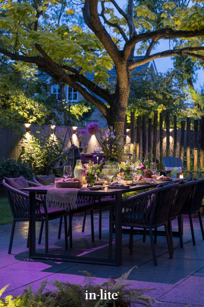 A close-up twilight view of an elegantly styled outdoor dining area set on premium paving. A vibrant purple light wash highlights the ground beneath the dark, woven dining chairs and table, which is dressed with floral arrangements and tableware. In the background, warm in-lite architectural wall lights create scalloped patterns along a timber fence, while a mature tree's canopy is softly uplit, demonstrating a layered, high-end low-voltage lighting design.