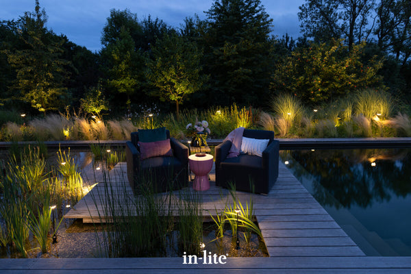 Atmospheric lighting by in-lite. SWAY 12v outdoor bollard light fixtures illuminating tall grasses, perennials in borders around a large waterside decking and outdoor seating space, illuminated with a SWAY Table, a rechargeable outdoor lamp lighting fixture.