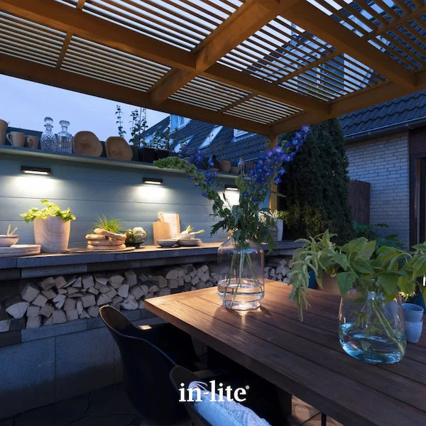 in-lite atmospheric garden lighting. in-lite WEDGE SLIM DARK 12v Outdoor Wall Lights positioned on wooden wall and shelving unit installed within a rustic style outdoor kitchen. Three slim wall fixtures are illuminating the wall and kitchen unit surfaces with an indirect, concentrated warm white light. The wall and kitchen unit features as a partition in an Alresco dining and outdoor living space covered by a slatted roof pergola.