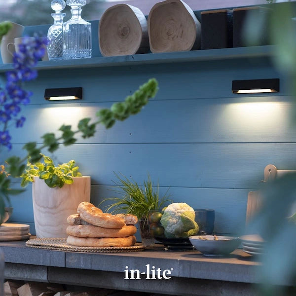 in-lite atmospheric garden lighting. in-lite WEDGE SLIM DARK 12v Outdoor Wall Lights positioned on wooden wall and shelving unit installed within a rustic style outdoor kitchen. The slim wall fixture is producing an indirect, concentrated warm white light, illuminating the wall and kitchen unit surfaces.